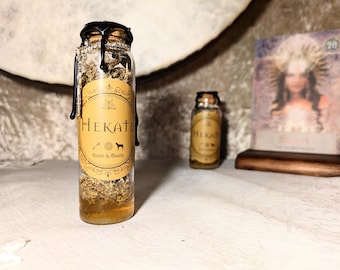 Könnte beinhalten: Eine Glasflasche mit Korken und einem Etikett mit der Aufschrift "Hekate". Die Flasche ist mit Kräutern und Flüssigkeit gefüllt. Der Verschluss ist mit schwarzem Wachs versiegelt. Eine kleinere Flasche und eine Tarotkarte befinden sich im Hintergrund.