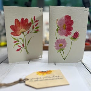 May include: Two watercolour floral art cards with scalloped edges. One card features a red flower with green leaves, the other has pink and purple flowers. A tag with a sunflower image and text is in front.