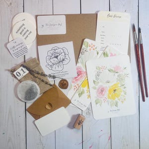May include: An assortment of stationery items, including cards with floral designs, a book review card, and a small envelope. There are also stickers with inspirational quotes, a tea bag, and paintbrushes. The items are arranged on a white wooden surface.