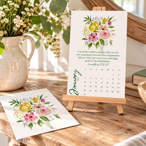 May include: A January 2027 calendar card on a small wooden easel, featuring a watercolor floral bouquet in yellow, pink, and green. The text reads: It is of the LORD's mercies that we are not consumed, because his compassions fail not.
