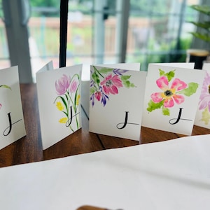 Può includere: Una collezione di biglietti di auguri floreali ad acquerello. Ogni biglietto presenta un diverso design floreale nei toni del rosa, viola e giallo, con foglie verdi. La lettera "J" è scritta in corsivo nero su ogni biglietto.