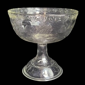 May include: A clear glass pedestal bowl with a decorative leaf pattern. The bowl has a wide, round base and a tall, slender stem.