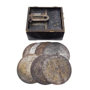 Puede incluir: Una caja de música antigua con una caja de madera oscura y un mecanismo de metal. Debajo de la caja hay varios discos metálicos circulares con patrones intrincados, probablemente para reproducir música. Los objetos parecen antiguos y bien conservados.