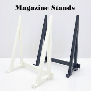 May include: Two magazine stands, one white and one black, are shown on a white surface. The stands are made of plastic and have a simple design. Both stands are labelled "PTP".