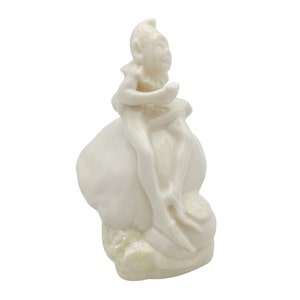 May include: White ceramic figurine of a fairy sitting on a mushroom. The fairy has wings and is holding a flower.