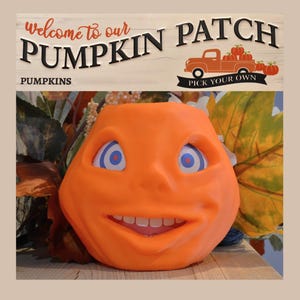 May include: An orange pumpkin-shaped ornament with a smiling face, blue eyes, and white teeth. The background features a sign that reads "Welcome to our Pumpkin Patch" and a truck carrying pumpkins. The sign also says "Pick Your Own".