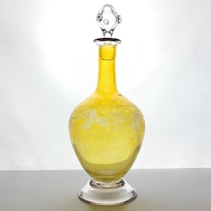 Puede incluir: Una decantador de vidrio amarillo con un tapón de vidrio transparente. La decantador tiene una superficie texturizada y un cuerpo redondo. El tapón tiene un diseño decorativo.