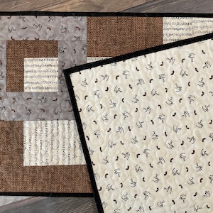 May include: Two brown and black quilted placemats with a coffee bean pattern. The placemats have a black border and a centre section with a burlap-like texture. The text "coffee, kaffe, kaffee, kaffekava, koffiekapekah" is printed on the placemats.