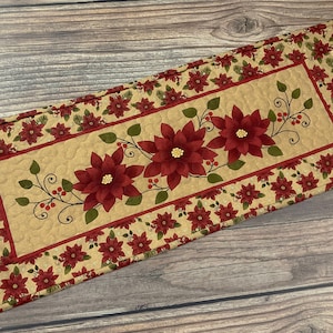 May include: A beige quilted table runner with a red and green poinsettia design. The runner has a border of red and green poinsettias and a center panel with a larger poinsettia design.