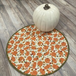 May include: A round table runner with a white background and a pattern of orange pumpkins and brown leaves. The runner has a green border.