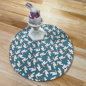 May include: A white ceramic bunny figurine with a pink waistcoat and an Easter egg sits on a white pedestal. The pedestal rests on a round teal placemat patterned with white bunnies wearing floral wreaths. The placemat is approx. 30 cm in diameter.