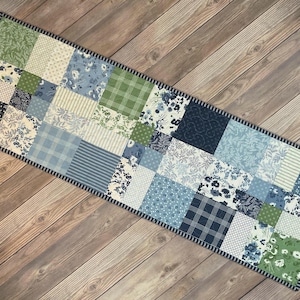 Puede incluir: Un camino de mesa rectangular de patchwork con una variedad de telas estampadas en azul, verde y crema. El camino de mesa está bordeado con una tela a rayas azul oscuro y blanco. El camino de mesa está colocado sobre una superficie de madera.
