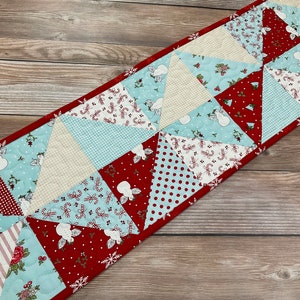 May include: A red, white, and blue quilted table runner with a snowflake pattern. The runner is made of various fabrics with different patterns, including polka dots, stripes, and candy canes.