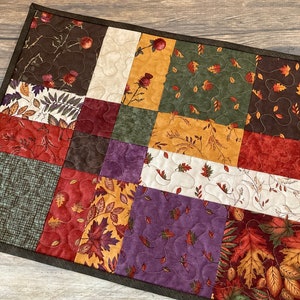 May include: A quilted table runner with a patchwork design featuring various autumn-themed fabrics in shades of brown, green, red, purple, and cream. The fabrics feature different patterns of leaves, branches, and floral designs.