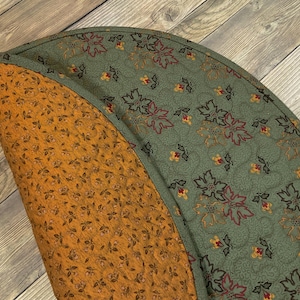 May include: A circular quilted fabric mat featuring a reversible design. One side displays a sage green background with a repeating pattern of dark red and gold leaves. The other side features a mustard yellow background with small brown floral sprigs.