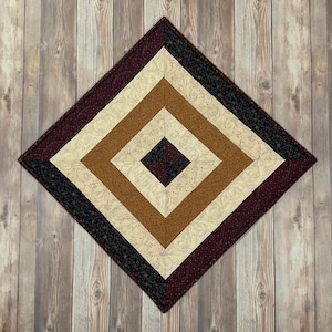 May include: A square quilted table runner with a geometric design. The runner features a burgundy, cream, and brown color scheme. The center of the runner is a dark burgundy square surrounded by a cream square, then a brown square, and finally a burgundy border.