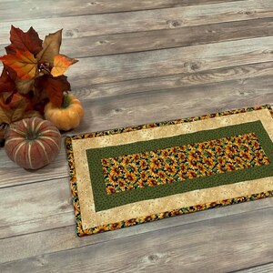 May include: A rectangular table runner with a green and beige border and a sunflower pattern in the center. The runner is on a wooden surface with two small pumpkins and fall leaves.