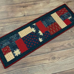 May include: A rectangular quilted table runner with a patriotic theme. The runner features blocks of red, navy blue, and tan fabric. Some blocks have red stars on a navy background, while others have a tan and red pattern. The edges are trimmed in red.
