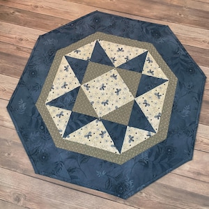 Può includere: Un runner da tavolo trapuntato ottagonale nei toni del blu, beige e crema. Il bordo esterno è una stampa floreale blu scuro, che incornicia un disegno centrale di triangoli e motivi floreali. Un elemento decorativo per un tavolo.