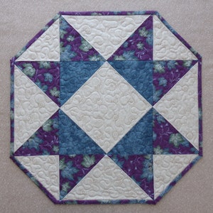 Puede incluir: Caminero de mesa acolchado octogonal con un diseño geométrico en tela blanca, morada y azul. El centro del camino de mesa tiene una tela blanca con un patrón de remolino.