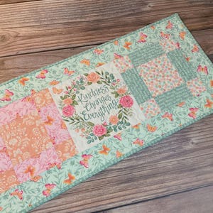 May include: A rectangular table runner with a patchwork design. The runner features squares of pink, peach, and floral patterns, with a central panel that reads "Kindness Changes Everything" surrounded by flowers. The border is light green with butterfly accents.