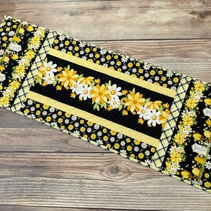 May include: A black and yellow table runner with a floral design featuring white daisies and yellow flowers. The runner has a black and white checkered border and a yellow and white polka dot border.