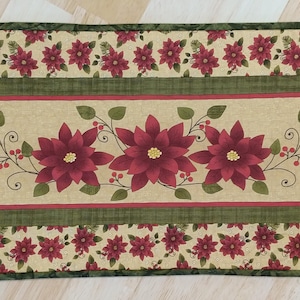 May include: A green and beige table runner with a red poinsettia design. The runner has a border of red poinsettias and green leaves.