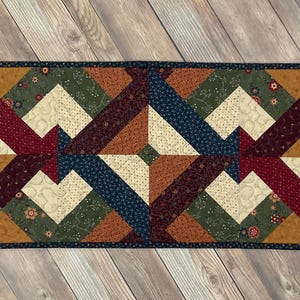 May include: A quilted table runner with a geometric pattern in shades of brown, red, green, and blue. The runner has a dark blue border.