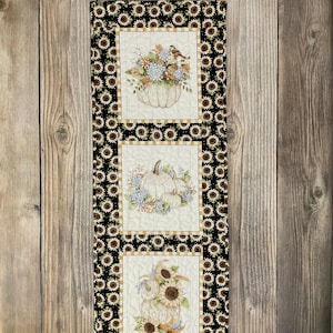 May include: A black and white quilted wall hanging with three panels featuring white pumpkins, sunflowers, and fall foliage. Each panel is framed with a black and yellow sunflower print border.