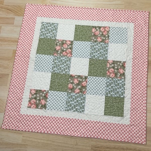 May include: A pink and white gingham border surrounds a patchwork quilt with a variety of floral and patterned fabric squares. The quilt is made up of 16 squares, each with a different design.