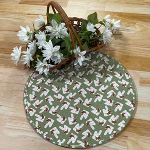 May include: A green round placemat with a white bunny rabbit pattern. The placemat has a white border and is made of fabric. A wicker basket filled with white flowers sits on top of the placemat.