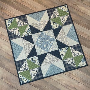 Puede incluir: Colcha cuadrada con un diseño de estrella. La colcha presenta un borde azul marino y un diseño central de estrella en tonos de azul, blanco y verde. La estrella está compuesta por varias telas con estampados florales y geométricos.