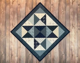 Puede incluir: Un edredón cuadrado con un diseño geométrico en tonos azul, crema y marino. El edredón presenta triángulos y cuadrados con varios patrones, incluyendo florales, cuadros y colores lisos. El edredón se exhibe sobre una superficie de madera.