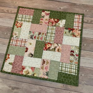 May include: A square patchwork quilt with a green border. The quilt features rectangular blocks of various fabrics, including floral, butterfly, and plaid patterns in shades of pink, green, and white. The quilt is approximately 12 inches square.