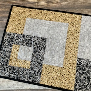 May include: A black and gold table runner with a geometric pattern. The runner is made of fabric with a quilted texture. The pattern features a combination of black, gold, and white fabrics.