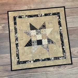 May include: A square quilted wall hanging featuring a star design. The star is composed of various patterned fabrics in black, white, and gray. The background is a beige fabric, and the border is black with a floral pattern. The piece is finished with a black binding.