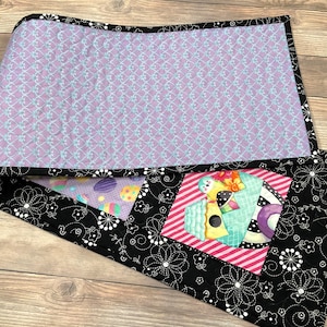 May include: A purple and black quilted table runner with a floral design and a birdhouse with eggs. The runner is approximately 14 inches wide and 36 inches long.