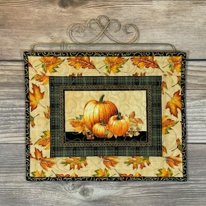 May include: A square quilted wall hanging featuring a central panel of orange pumpkins on a cream background, surrounded by a dark green plaid border and an outer border of orange and yellow autumn leaves, suspended from a decorative metal hanger.