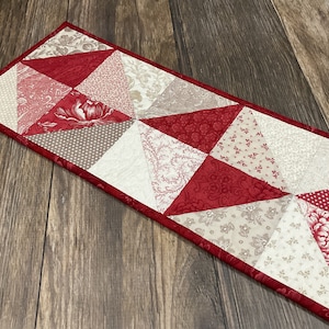 May include: A red and cream quilted table runner with a geometric pattern of triangles. The runner has a red border.