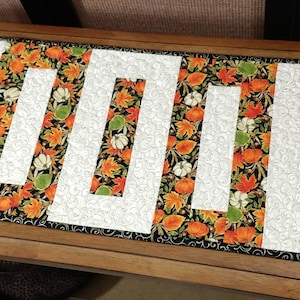 May include: A table runner with a fall-themed fabric featuring orange, green, and white leaves on a black background. The runner has five white rectangular cutouts.