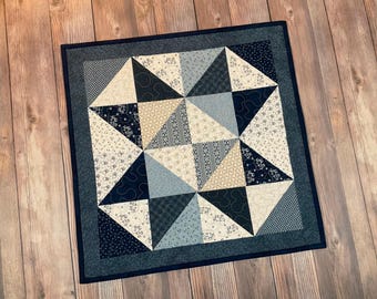 Puede incluir: Un edredón cuadrado con un diseño geométrico de estrella. El edredón presenta triángulos en tonos de azul, crema y beige, con varios patrones. Un borde azul oscuro enmarca el edredón, que se muestra sobre una superficie de madera.