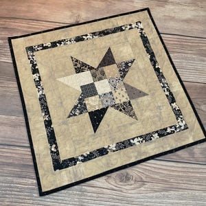 May include: A square quilted wall hanging with a star design. The star is composed of various patterned fabrics in shades of beige, black, and gray. The star is centered on a beige background, framed by a floral border and a black edge.
