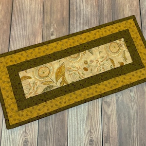 May include: Rectangular quilted table runner with a floral design. The runner features a central panel with beige fabric and botanical prints, framed by olive green and gold borders. The overall design is rectangular.