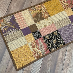Könnte beinhalten: Ein rechteckiger Patchwork-Tischläufer mit einer Vielzahl von Stoffquadraten in Senfgelb, Dunkelviolett, Altrosa, Creme und Schwarz. Die Stoffquadrate zeigen verschiedene Blumen-, Karomuster und geometrische Designs mit einer braunen Umrandung.