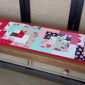 May include: A patchwork table runner with a red, white, and blue color scheme. The runner is made of various floral and geometric fabrics.