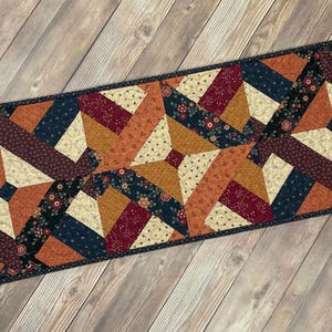 May include: A quilted table runner with a geometric pattern in shades of brown, orange, cream, navy blue, and burgundy. The runner is made of fabric with a floral print.