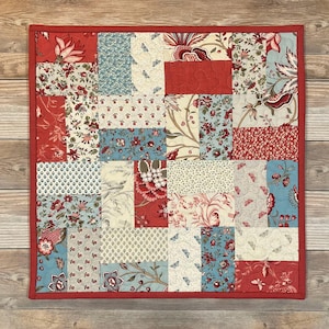 May include: A square patchwork quilt with a red border. The quilt is made of various fabric squares in red, cream, and blue with floral and patterned designs. The quilt is approximately 12 inches by 12 inches.
