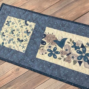 May include: A blue and cream quilted table runner with a floral and butterfly design. The runner features a border of blue fabric with a white floral pattern. The center of the runner features a cream fabric with a floral and butterfly design. The text "Butterflies" is repeated throughout the design.
