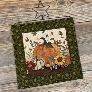 May include: A square fabric wall hanging featuring a central panel with a large orange pumpkin, a smaller white pumpkin, a sunflower, and autumn leaves. The border is dark green with a leaf pattern, and a black wire star hanger is attached at the top.