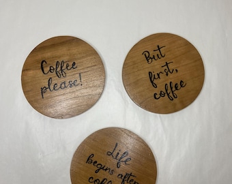 May include: Three round wooden coasters with black lettering. The coasters feature the following text: "Coffee please!" "But first, coffee" and "Life begins after coffee!"